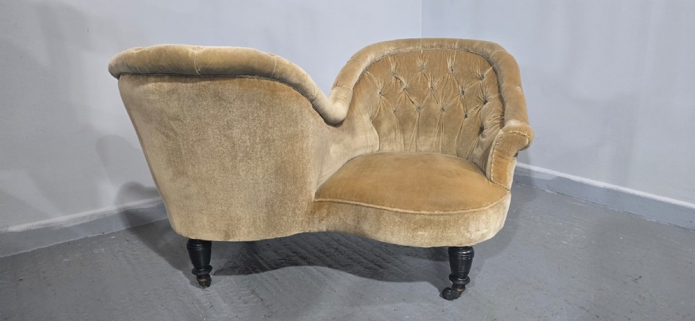 An S-shaped Victorian conversation sofa upholstered in warm gold velvet with deep-buttoned backrests and turned dark timber legs on castors. View V