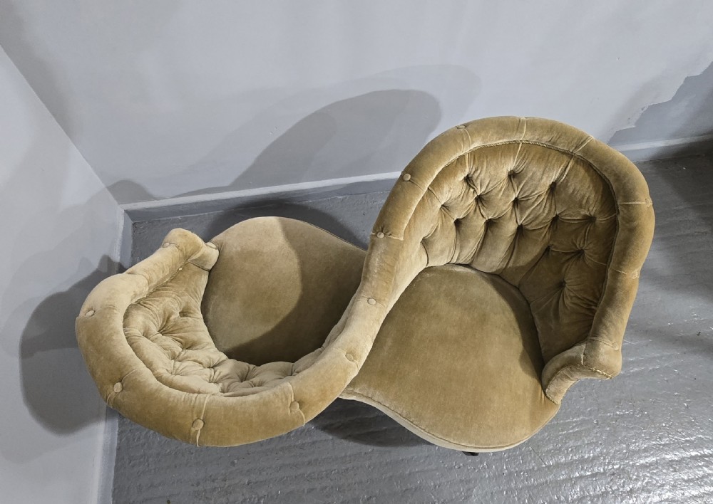 An S-shaped Victorian conversation sofa upholstered in warm gold velvet with deep-buttoned backrests and turned dark timber legs on castors. View VIII
