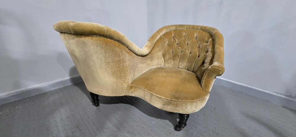 An S-shaped Victorian conversation sofa upholstered in warm gold velvet with deep-buttoned backrests and turned dark timber legs on castors. View XII