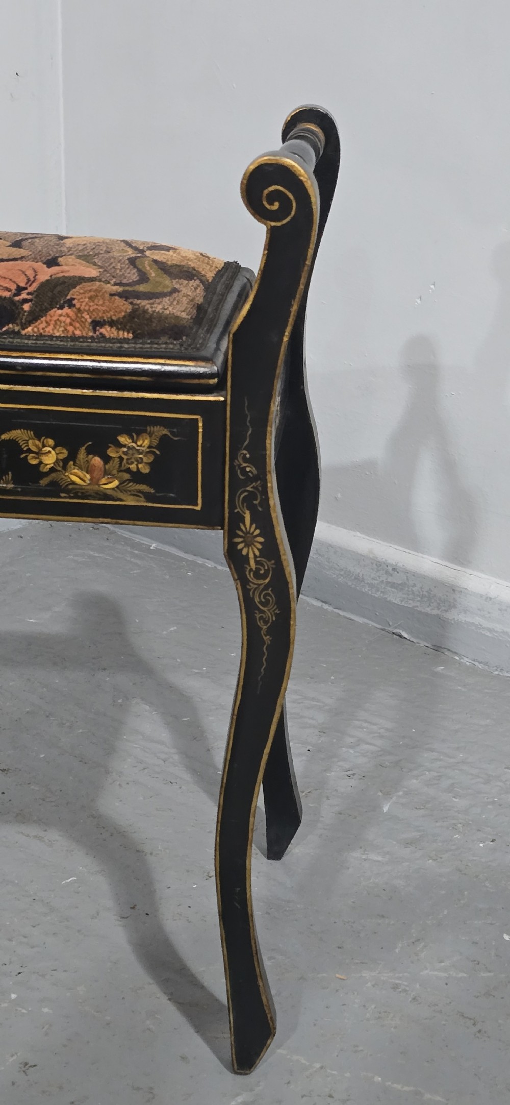 A black lacquered piano stool with hand-painted gold Chinoiserie designs, turned side handles, curved legs, and a floral tapestry seat. View IV