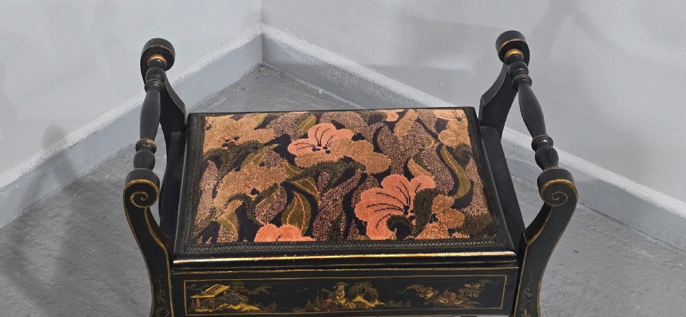 A black lacquered piano stool with hand-painted gold Chinoiserie designs, turned side handles, curved legs, and a floral tapestry seat. View V