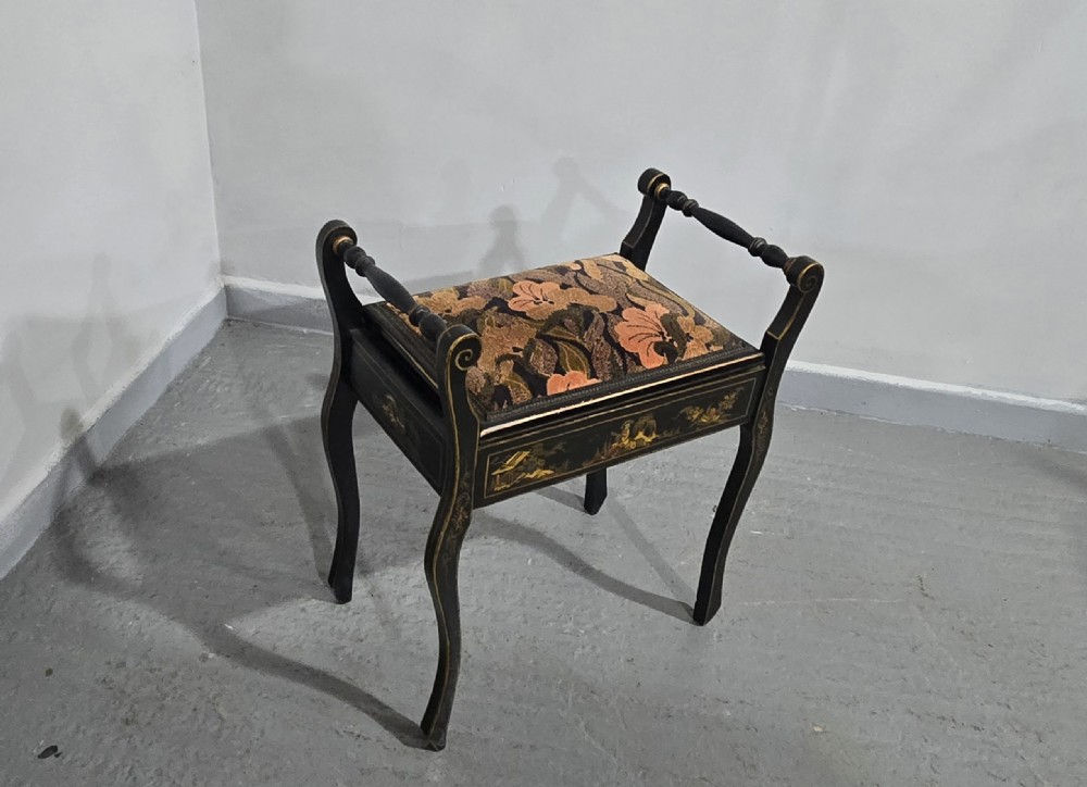 A black lacquered piano stool with hand-painted gold Chinoiserie designs, turned side handles, curved legs, and a floral tapestry seat. View VI