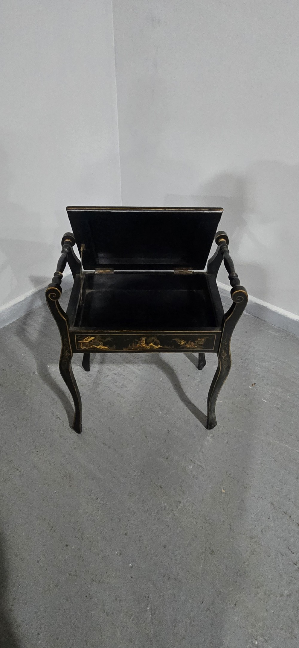 A black lacquered piano stool with hand-painted gold Chinoiserie designs, turned side handles, curved legs, and a floral tapestry seat. View VII
