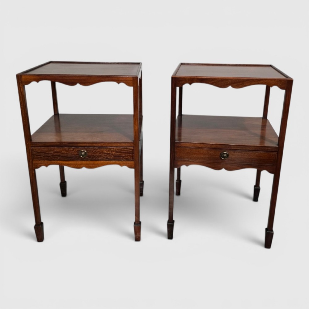 A pair of Georgian mahogany lamp tables featuring tray top rims, middle open shelves, single drawers with brass knobs, and shaped aprons.