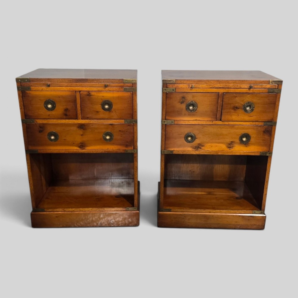 A pair of warm brown yew wood campaign style bedside tables featuring brass corner mounts, flush brass pull handles, drawers, and lower open shelves.