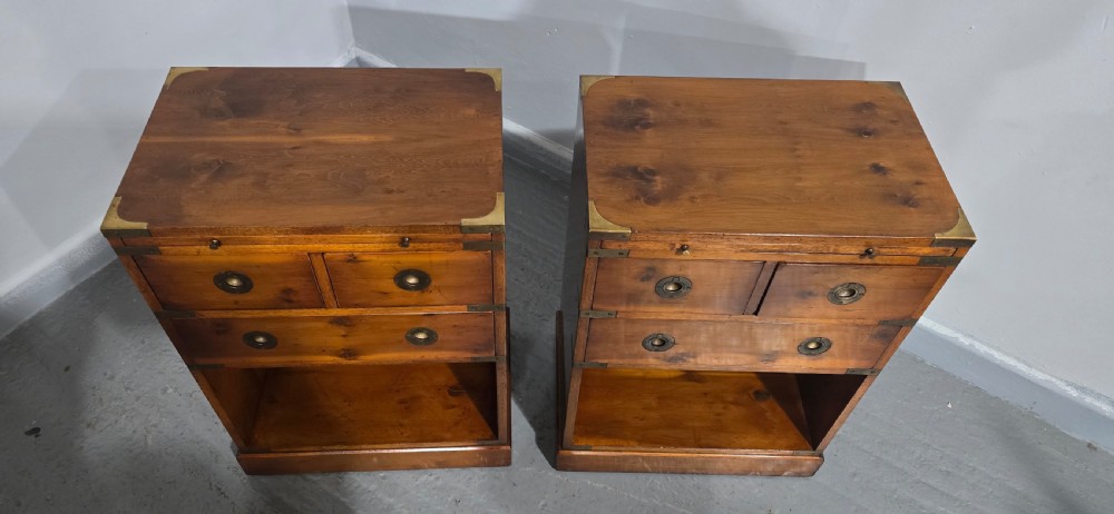 A pair of warm brown yew wood campaign style bedside tables featuring brass corner mounts, flush brass pull handles, drawers, and lower open shelves. View IV