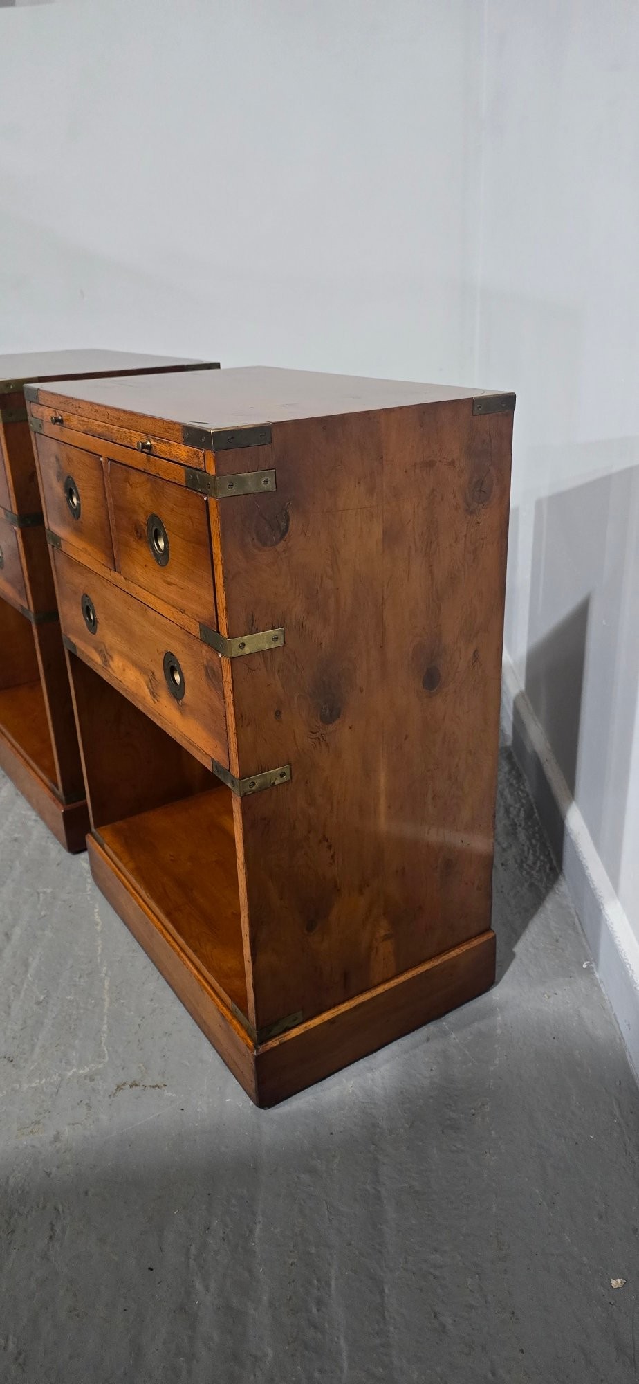 A pair of warm brown yew wood campaign style bedside tables featuring brass corner mounts, flush brass pull handles, drawers, and lower open shelves. View VI