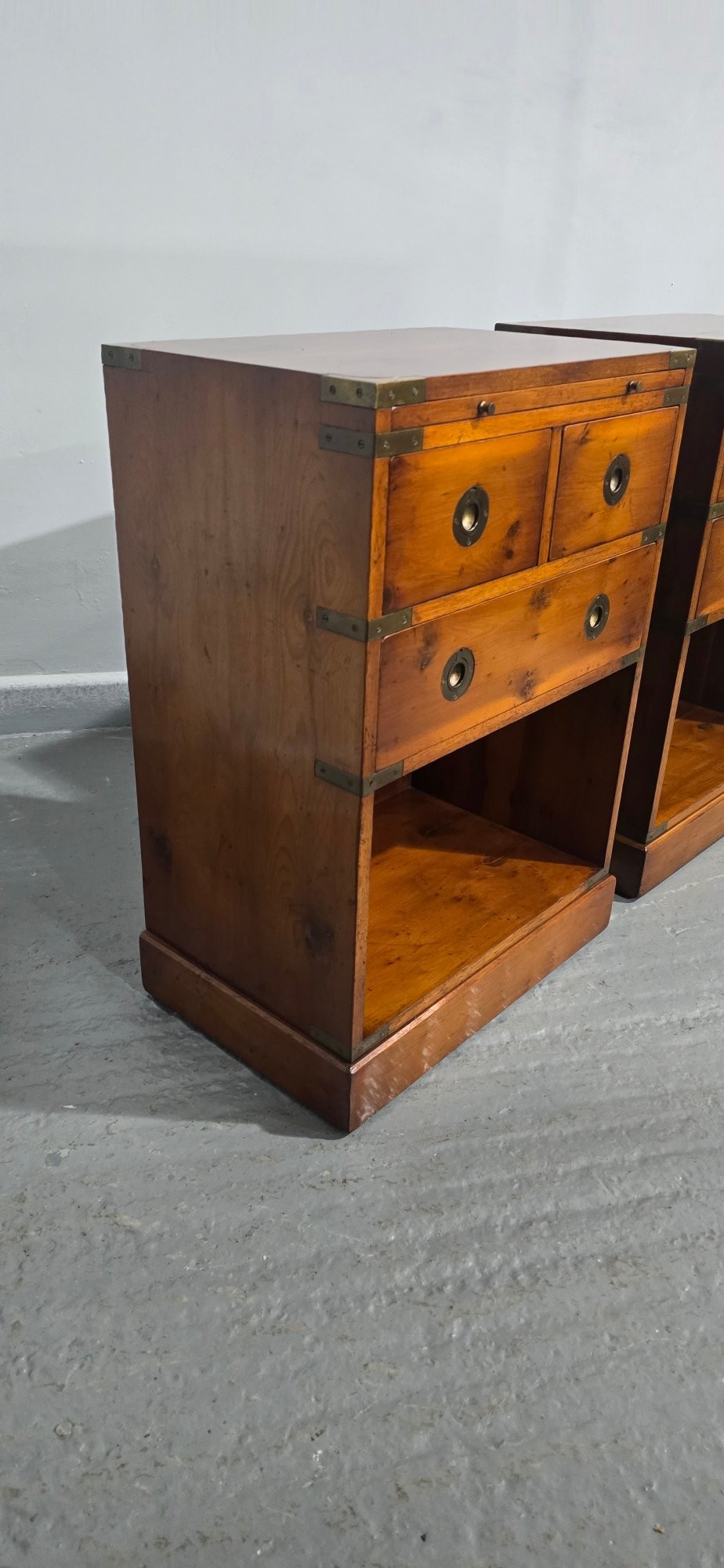 A pair of warm brown yew wood campaign style bedside tables featuring brass corner mounts, flush brass pull handles, drawers, and lower open shelves. View VII