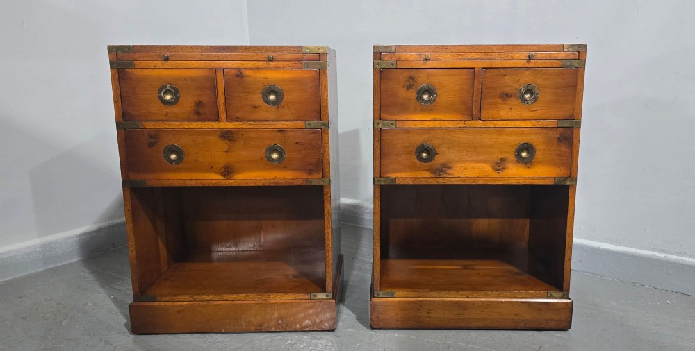 A pair of warm brown yew wood campaign style bedside tables featuring brass corner mounts, flush brass pull handles, drawers, and lower open shelves. View X