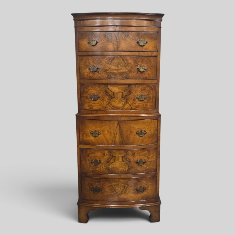 A small bowfront burr walnut chest on chest featuring stacked drawers with brass handles and bracket feet.