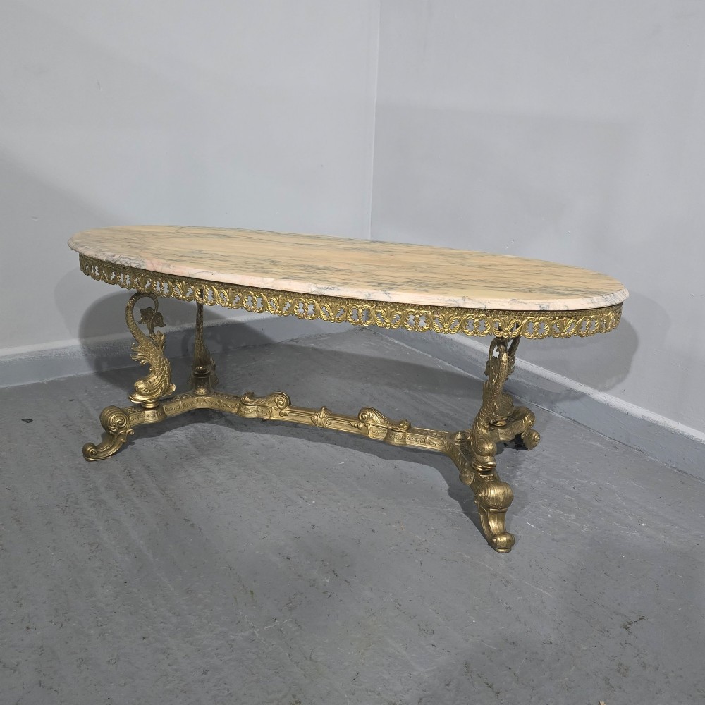 An oval gilt brass coffee table with an ornate pierced gallery apron, scrolled mythical legs, a central stretcher, and a polished veined pink marble top. View II