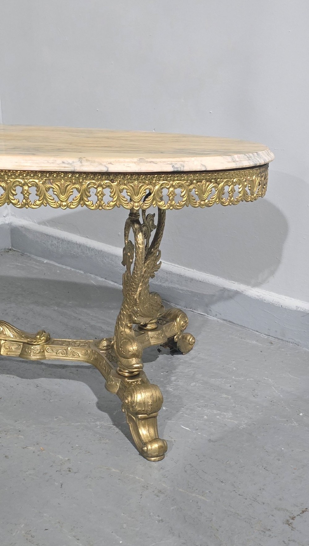 An oval gilt brass coffee table with an ornate pierced gallery apron, scrolled mythical legs, a central stretcher, and a polished veined pink marble top. View V