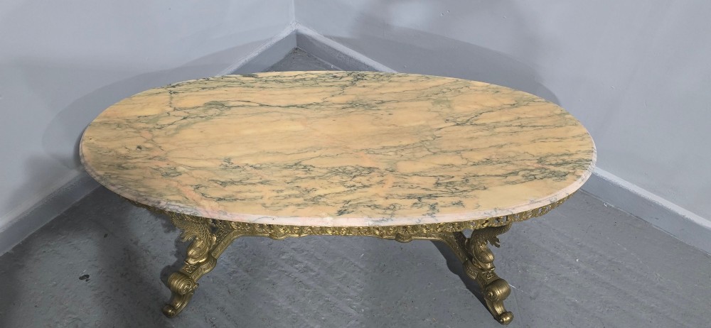 An oval gilt brass coffee table with an ornate pierced gallery apron, scrolled mythical legs, a central stretcher, and a polished veined pink marble top. View VII