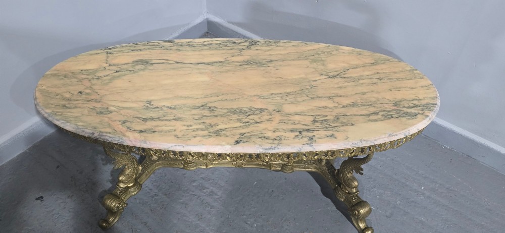 An oval gilt brass coffee table with an ornate pierced gallery apron, scrolled mythical legs, a central stretcher, and a polished veined pink marble top. View IX