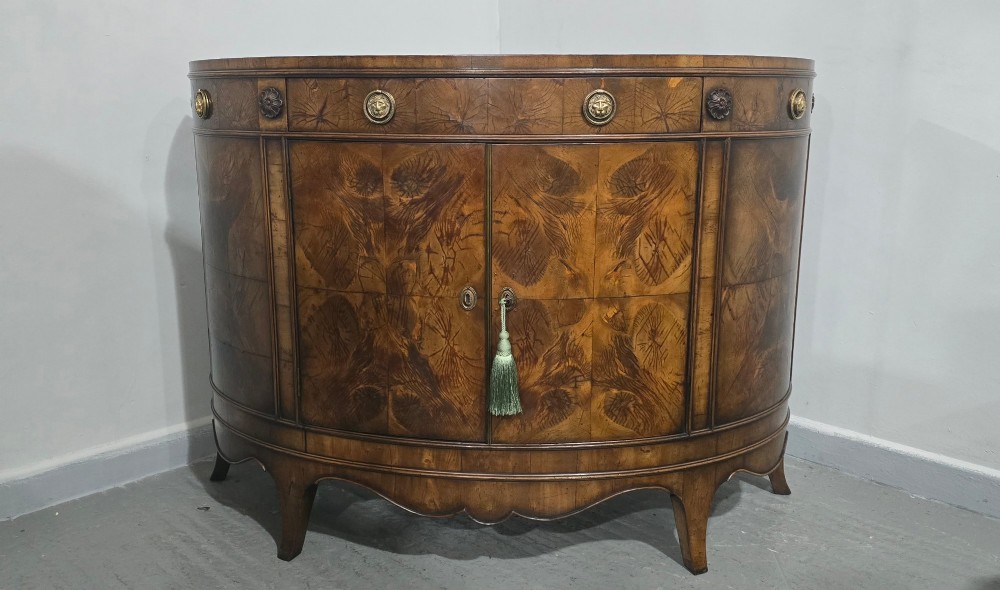 A semi-circular burr yew wood side cabinet featuring brass mounts along the frieze and central double doors. View II