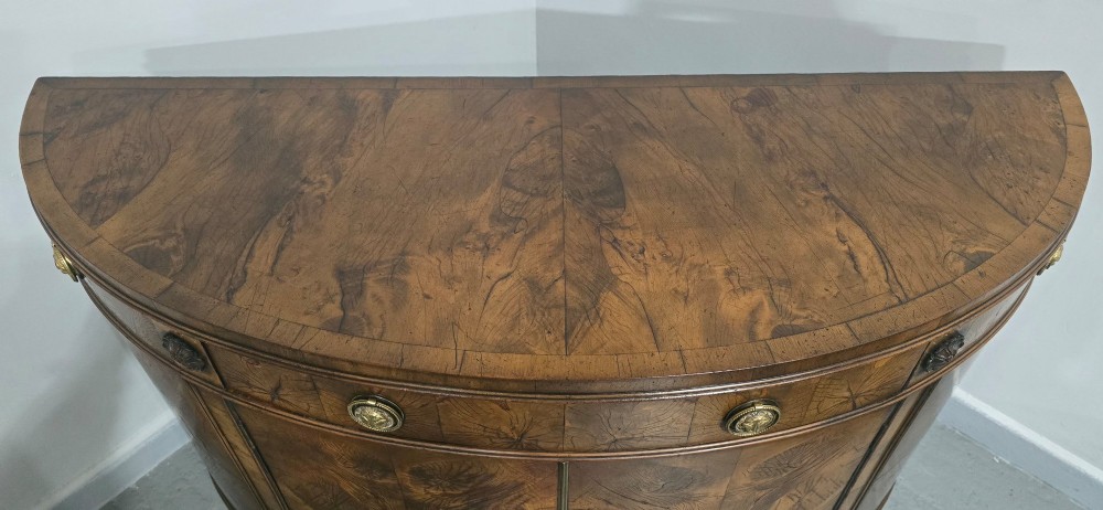 A semi-circular burr yew wood side cabinet featuring brass mounts along the frieze and central double doors. View VIII