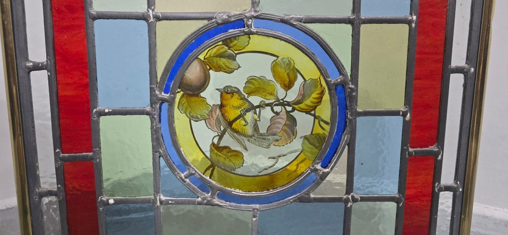A brass fire screen featuring a leaded stained glass panel with a central hand-painted bird roundel set on splayed brass feet. View II
