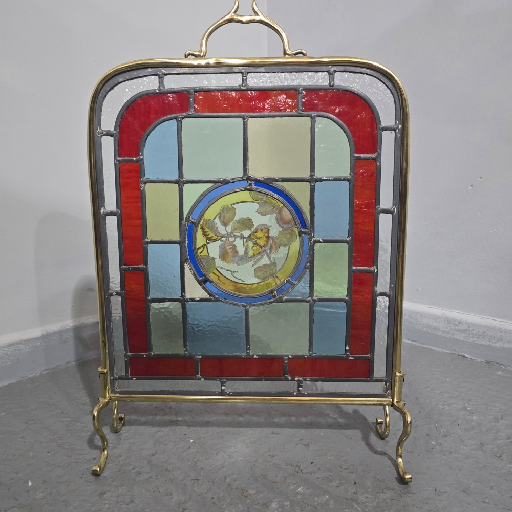 A brass fire screen featuring a leaded stained glass panel with a central hand-painted bird roundel set on splayed brass feet. View V