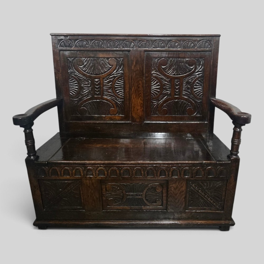 A dark carved oak monks bench featuring relief-carved panel work on the backrest and base, complete with curved armrests and a hinged storage seat.