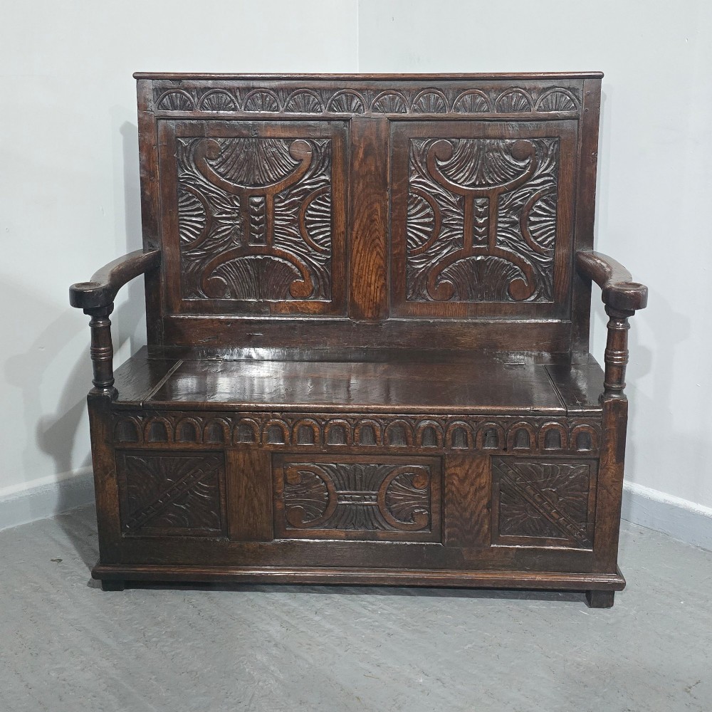 A dark carved oak monks bench featuring relief-carved panel work on the backrest and base, complete with curved armrests and a hinged storage seat. View II