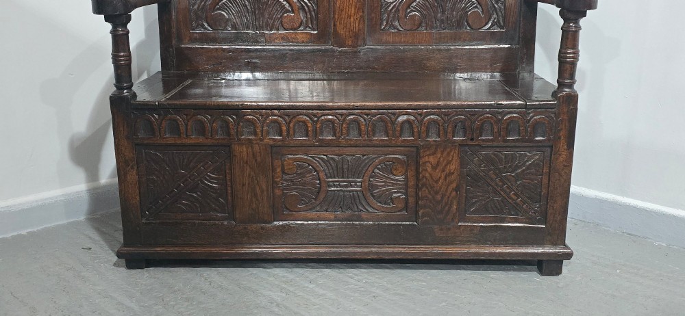 A dark carved oak monks bench featuring relief-carved panel work on the backrest and base, complete with curved armrests and a hinged storage seat. View III