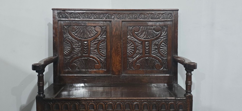 A dark carved oak monks bench featuring relief-carved panel work on the backrest and base, complete with curved armrests and a hinged storage seat. View IV