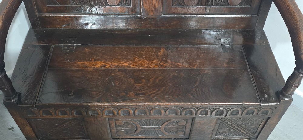 A dark carved oak monks bench featuring relief-carved panel work on the backrest and base, complete with curved armrests and a hinged storage seat. View V
