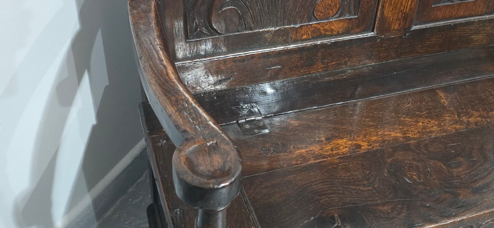 A dark carved oak monks bench featuring relief-carved panel work on the backrest and base, complete with curved armrests and a hinged storage seat. View VII