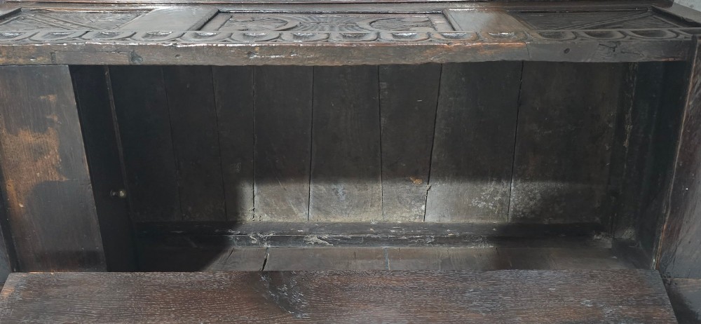 A dark carved oak monks bench featuring relief-carved panel work on the backrest and base, complete with curved armrests and a hinged storage seat. View X