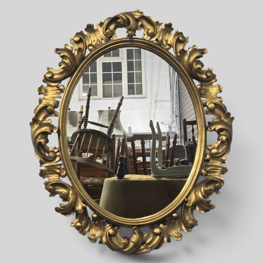 An oval wall mirror enclosed in a deeply carved gold giltwood frame featuring ornate acanthus leaf and scrolling foliage details.