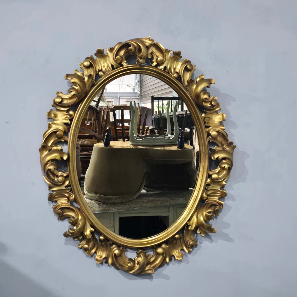 An oval wall mirror enclosed in a deeply carved gold giltwood frame featuring ornate acanthus leaf and scrolling foliage details. View II