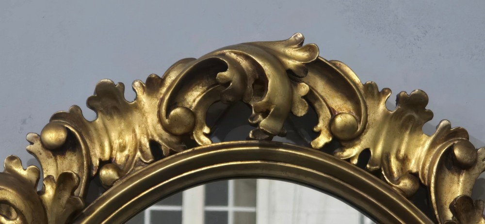 An oval wall mirror enclosed in a deeply carved gold giltwood frame featuring ornate acanthus leaf and scrolling foliage details. View V