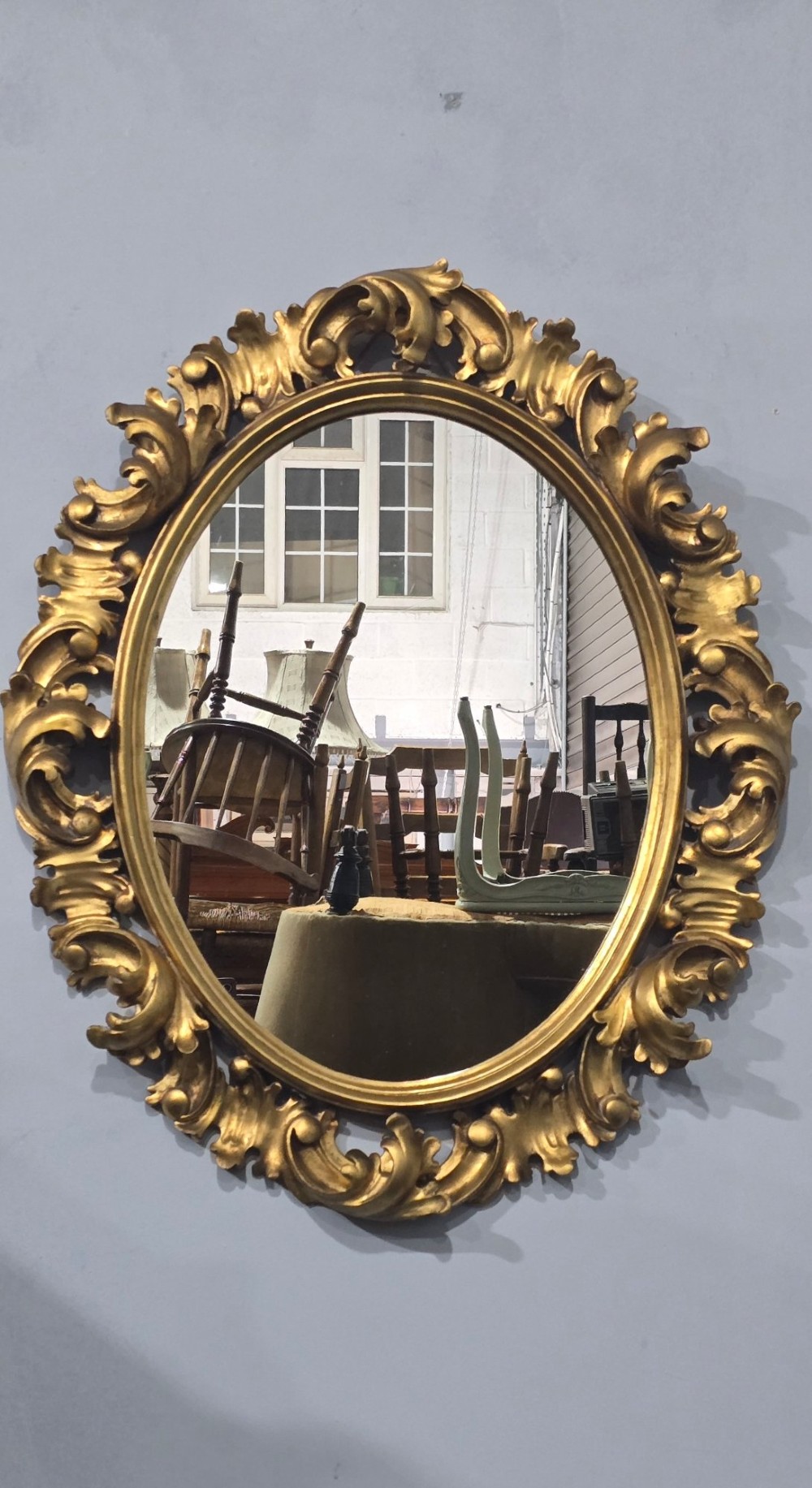 An oval wall mirror enclosed in a deeply carved gold giltwood frame featuring ornate acanthus leaf and scrolling foliage details. View VII
