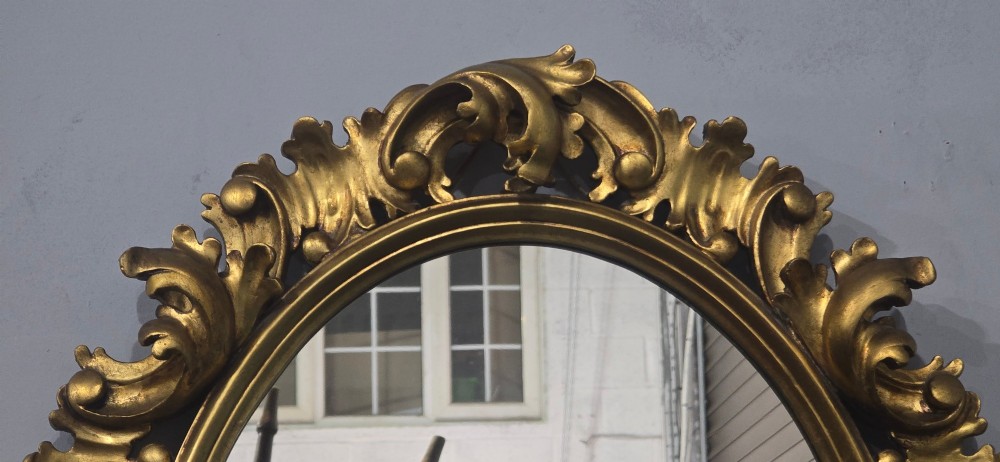 An oval wall mirror enclosed in a deeply carved gold giltwood frame featuring ornate acanthus leaf and scrolling foliage details. View X
