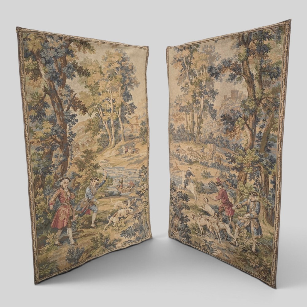 A pair of vertical woven French tapestries depicting men and hounds hunting within a dense green forest and natural landscape.