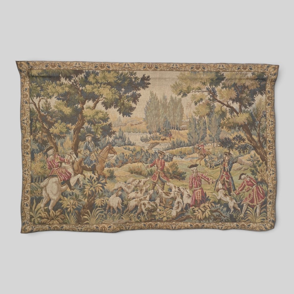 A large woven textile wall hanging featuring a French hunting scene with figures on horseback, hounds, and a forest landscape surrounded by a patterned border.