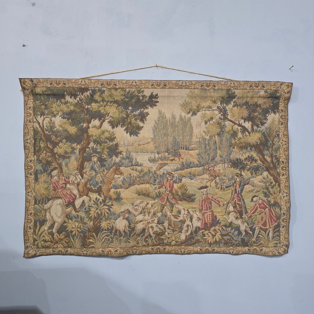 A large woven textile wall hanging featuring a French hunting scene with figures on horseback, hounds, and a forest landscape surrounded by a patterned border. View II