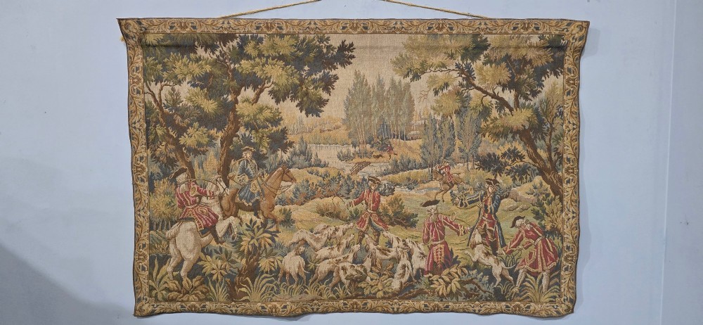 A large woven textile wall hanging featuring a French hunting scene with figures on horseback, hounds, and a forest landscape surrounded by a patterned border. View III