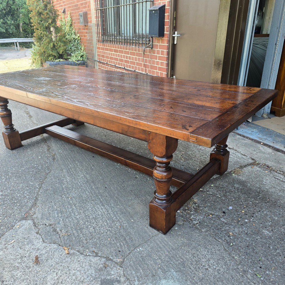 A large solid oak refectory dining table featuring a wide plank top, breadboard ends, turned baluster legs, and a low H-stretcher base. View XIV