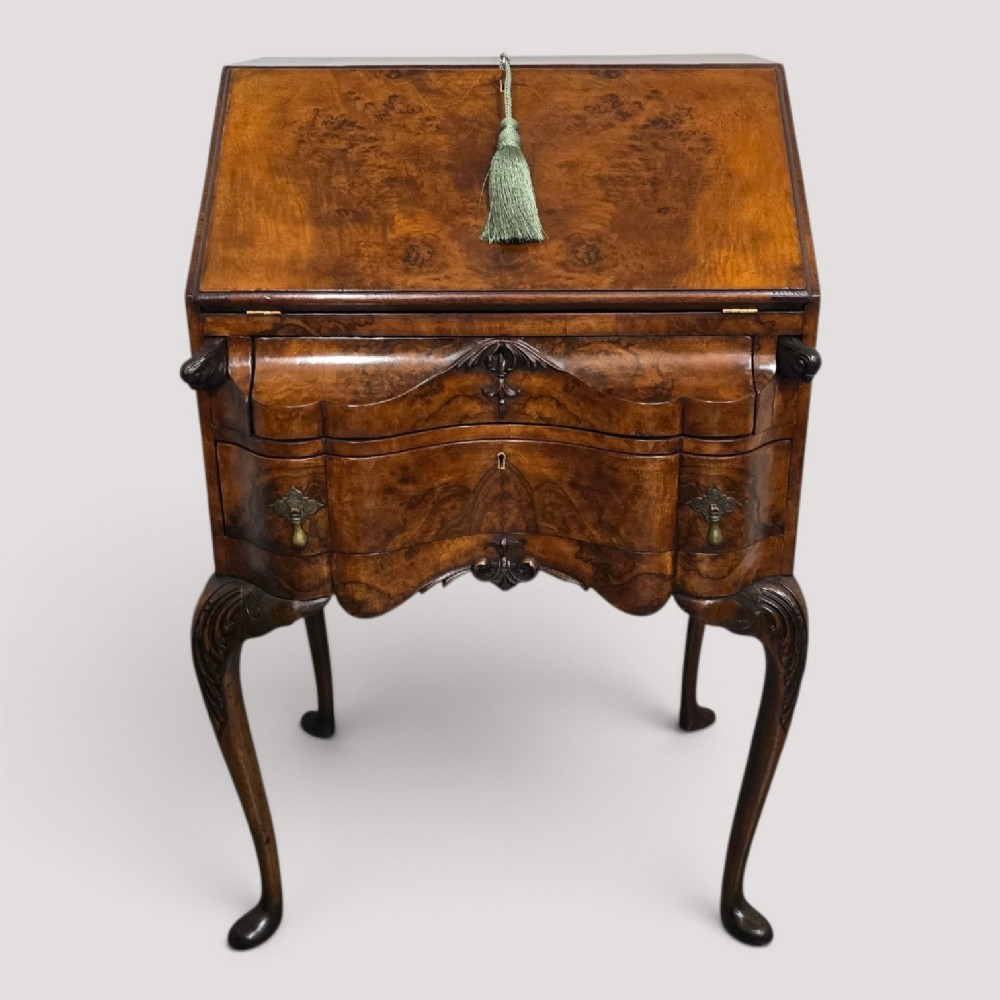 A Queen Anne style burr walnut writing bureau featuring a sloped front lid, shaped drawers with brass drop handles, and tall cabriole legs.