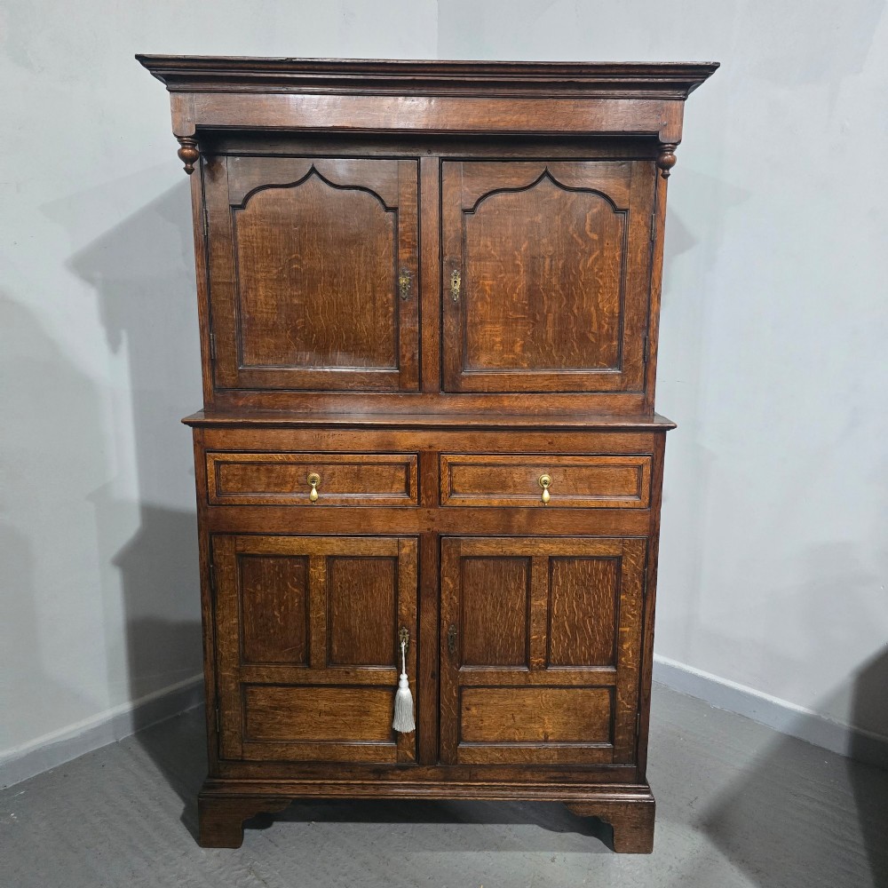 A dark oak Georgian court cupboard featuring an upper cabinet with arched panel doors, two central drawers, and a lower double-door cupboard on bracket feet. View XV