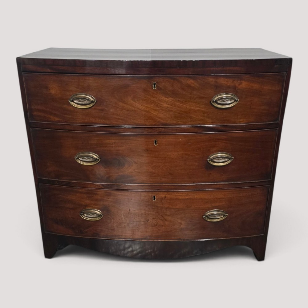 An antique Georgian mahogany bowfront chest featuring three drawers with oval brass handles and keyholes.