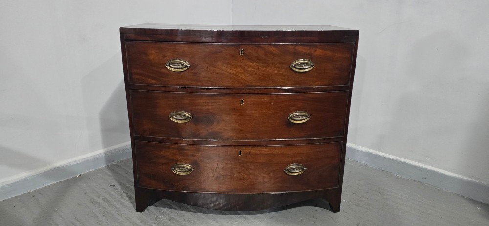 An antique Georgian mahogany bowfront chest featuring three drawers with oval brass handles and keyholes. View IV