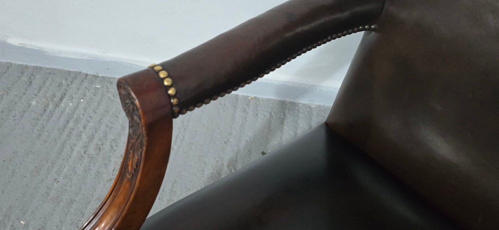 A dark brown leather upholstered swivel desk chair featuring carved mahogany armrests, brass studding, and splayed legs with brass castors. View VI