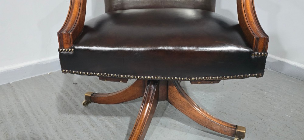 A dark brown leather upholstered swivel desk chair featuring carved mahogany armrests, brass studding, and splayed legs with brass castors. View VIII