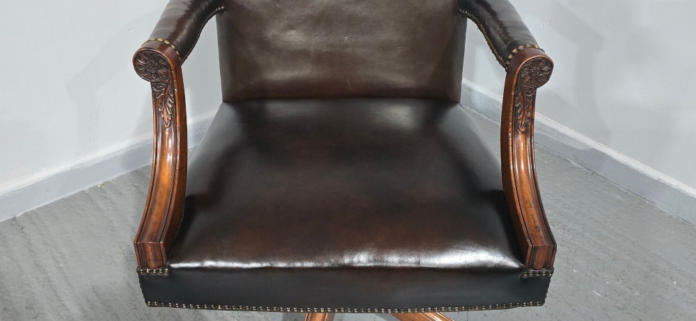 A dark brown leather upholstered swivel desk chair featuring carved mahogany armrests, brass studding, and splayed legs with brass castors. View IX