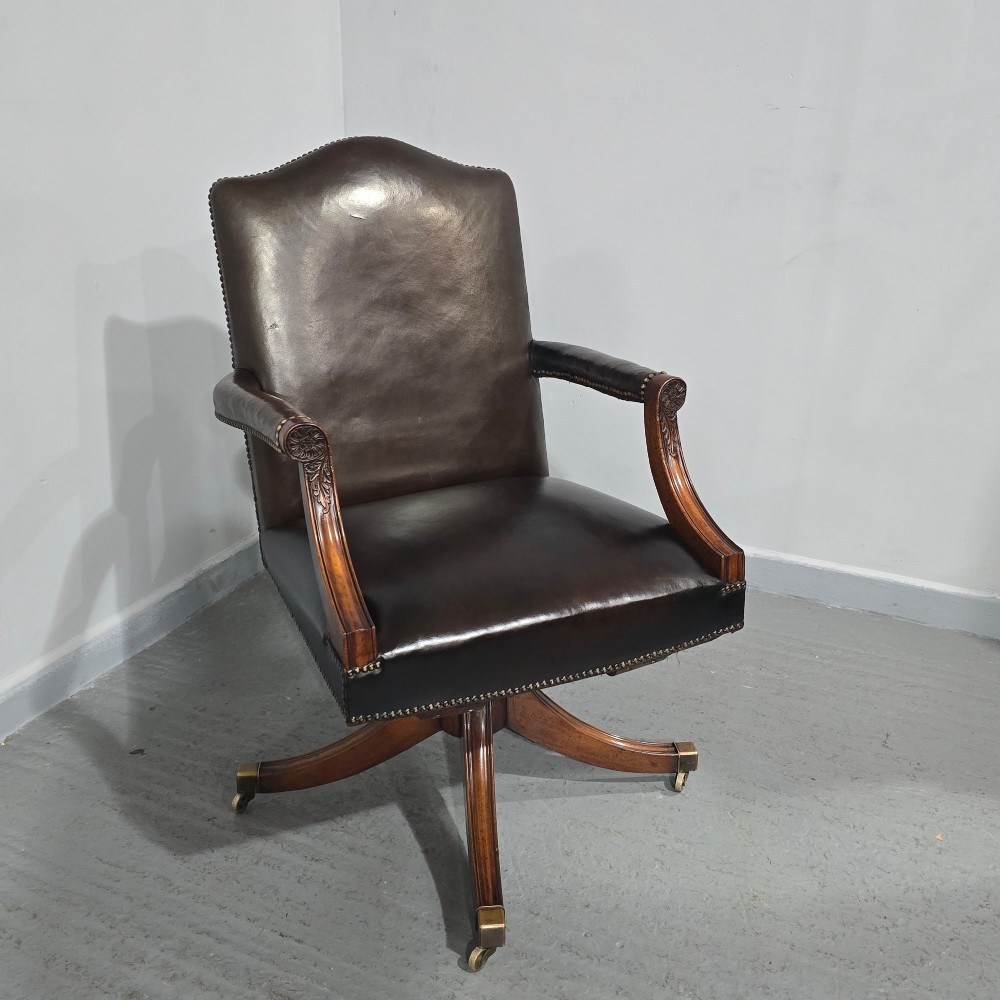 A dark brown leather upholstered swivel desk chair featuring carved mahogany armrests, brass studding, and splayed legs with brass castors. View XI