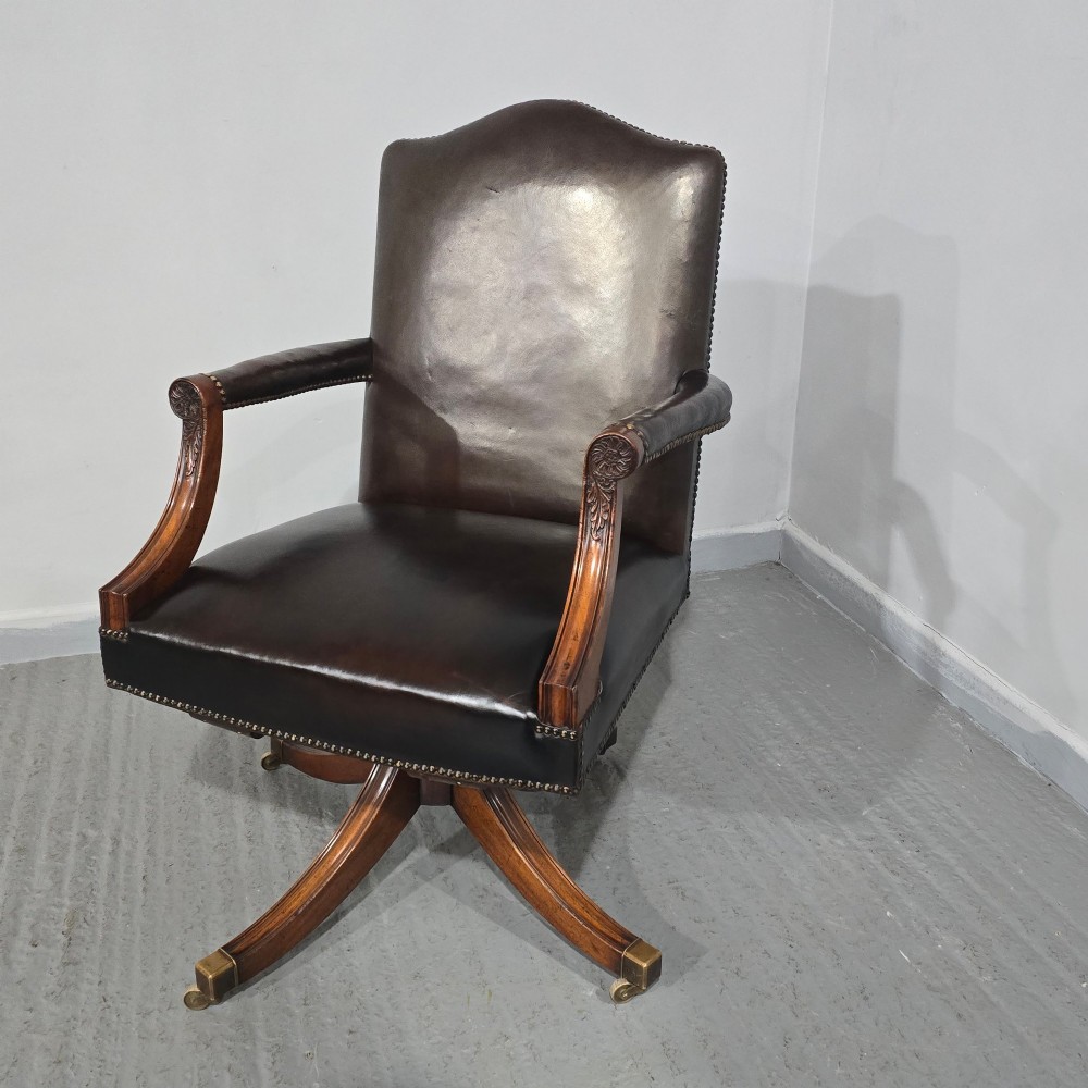 A dark brown leather upholstered swivel desk chair featuring carved mahogany armrests, brass studding, and splayed legs with brass castors. View XII