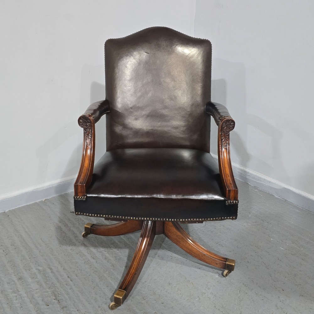 A dark brown leather upholstered swivel desk chair featuring carved mahogany armrests, brass studding, and splayed legs with brass castors. View XIII