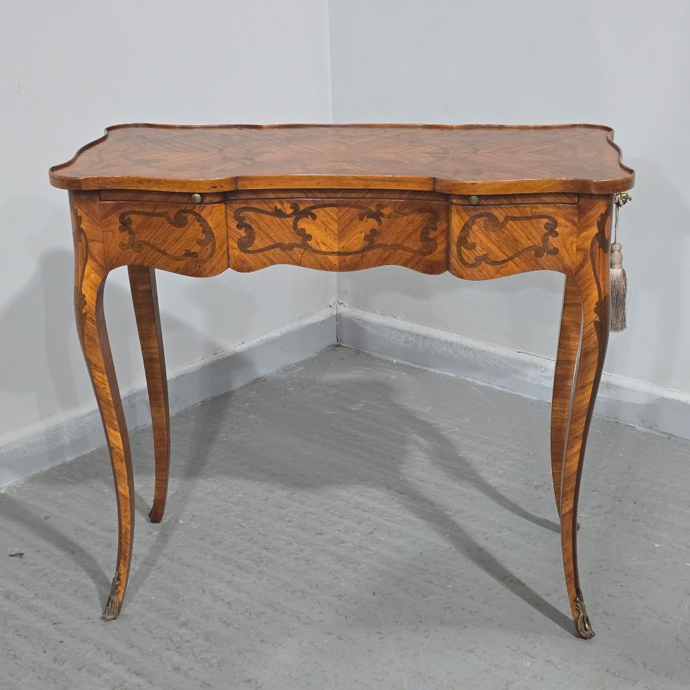 A small French Louis XV style writing table crafted from warm brown wood with scrolling marquetry, a serpentine edge, and slim cabriole legs. View III